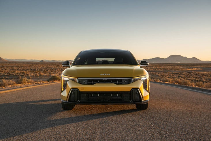 Rear three-quarter view of a light gray Kia EV6 driving away on a winding desert highway at sunset.
