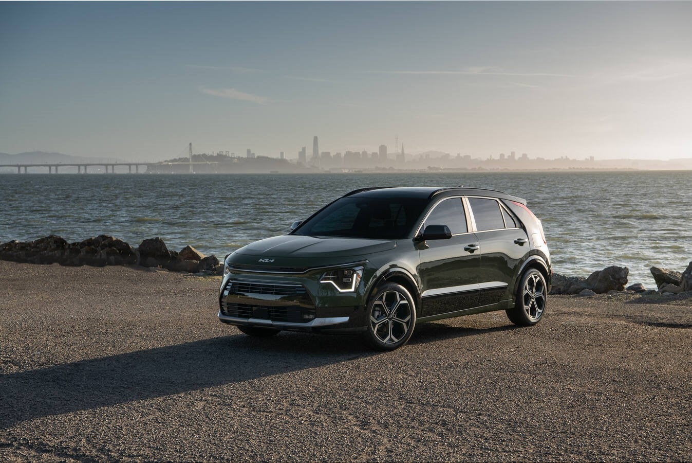 A dark green new Kia Niro in Phoenix is parked facing the viewer to the left on a paved lot in front of a large body of water with a large bridge and a city skyline barely visible far in the background.
