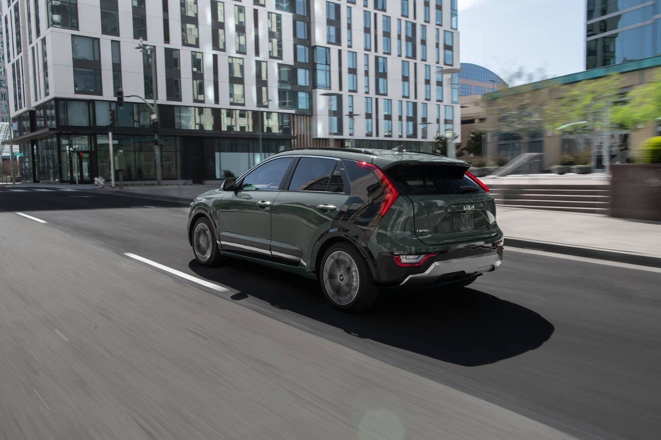 A dark green new Kia Niro in Peoria drives away from the viewer to the left on a city street with modern buildings in the background.