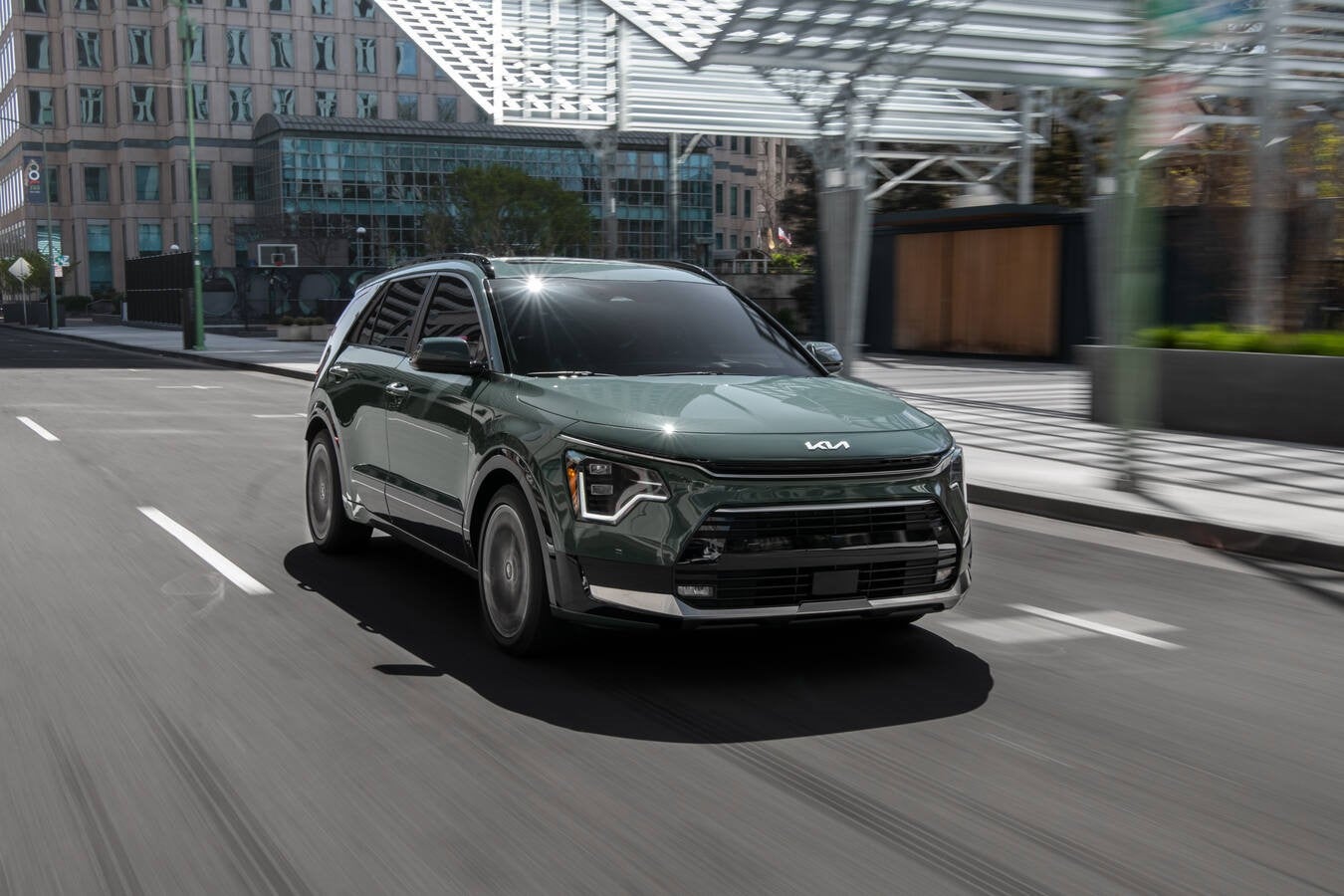 A dark green 2026 Kia Niro is driving toward the viewer to the righ in Phoenix on a busy city street under heavy sunlight.
