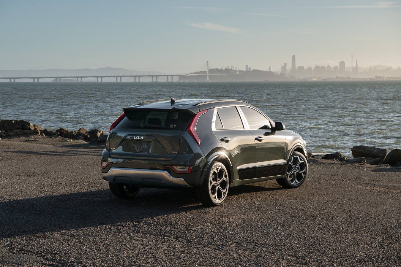 A dark green 2026 Kia Niro in Glendale is parked facing away from the viewer to the right on a paved lot in front of a large body of water with a large bridge and a city skyline barely visible far in the background.