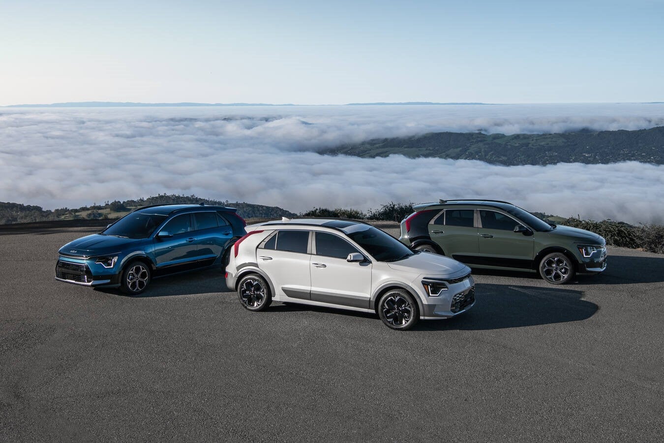 Three 2026 Kia Niro's (blue, left; white, center; dark green, right) are parked on a paved overlook on a mountain road with a valley filled with rolling clouds in the background.