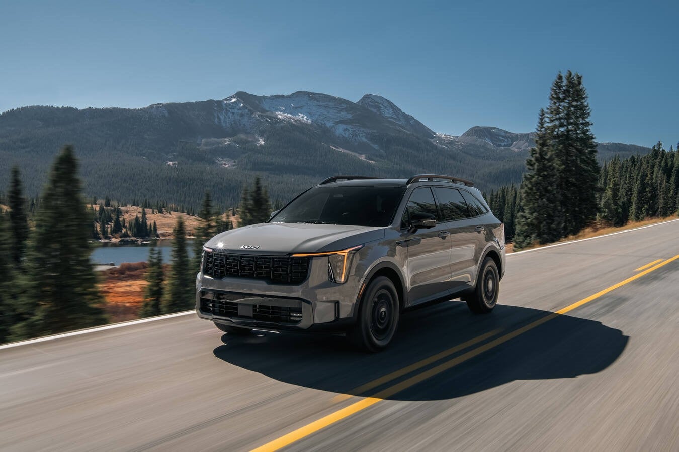 A grey 2026 Kia Sorento in Phoenix drives towards the viewer to the left on a two-lane road amongst tree-covered mountains.