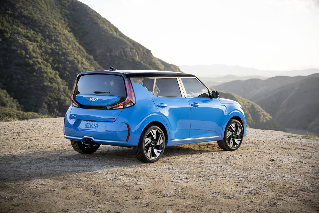 A blue new 2026 Kia Soul in Peoria is parked facing away from the viewer to the right on a gravel trail high in the mountains looking out into a lush valley.
