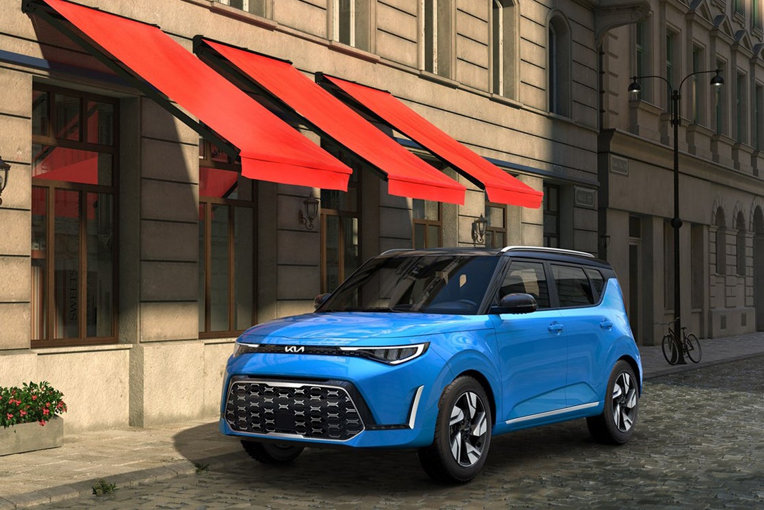 A blue new Soul in Phoenix is parked facing the viewer to the left on a quaint cobblestone city street next to a tan building facade with red overhangs.