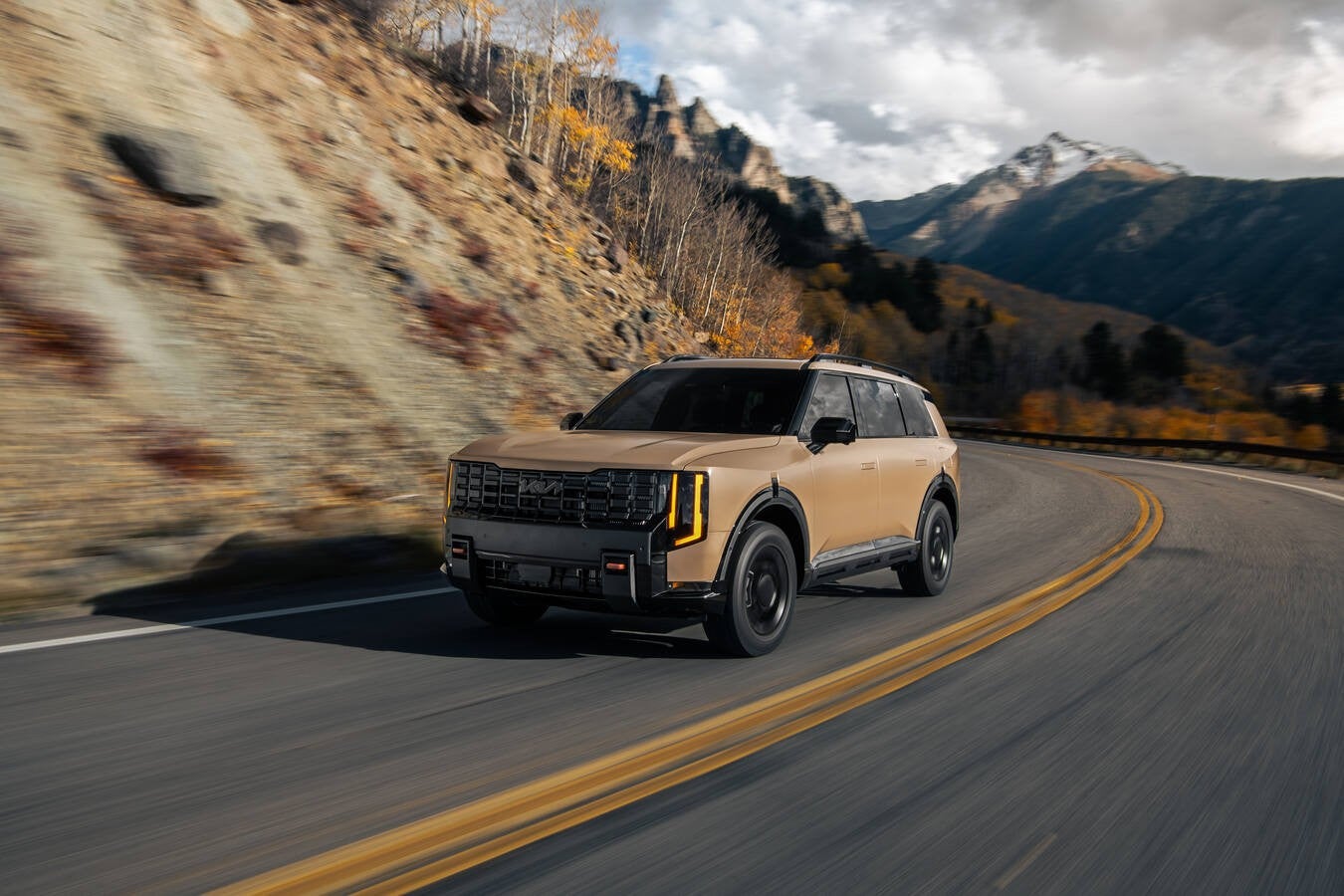 A brown 2026 Kia Telluride in Peoria is driving towards the viewer to the left on a two-lane mountain road on a cloudy day.