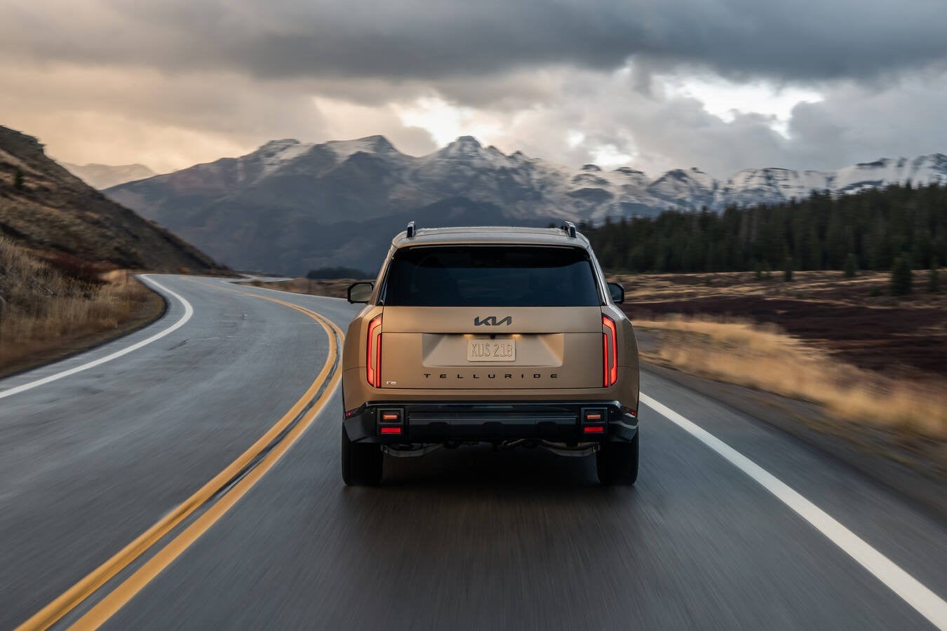 A brown new Kia Telluride in Phoenix is driving directly away from the viewer on a two-lane mountain road on a cloudy day.