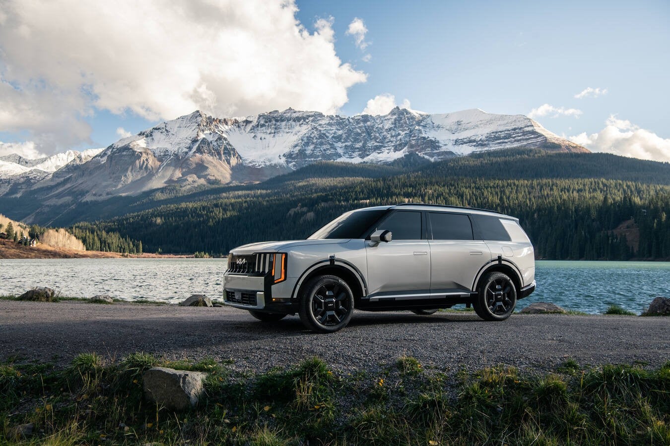 A silver new 2026 Kia Telluride Hybrid is parked facing left on a gravel lot next to a serene lake in front of a snowy mountain covered in trees on a sunny day.