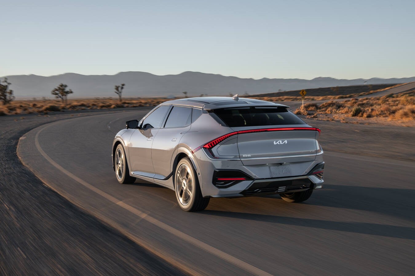 A white Kia EV9 electric SUV drives at high speed on a paved highway through a desert landscape.