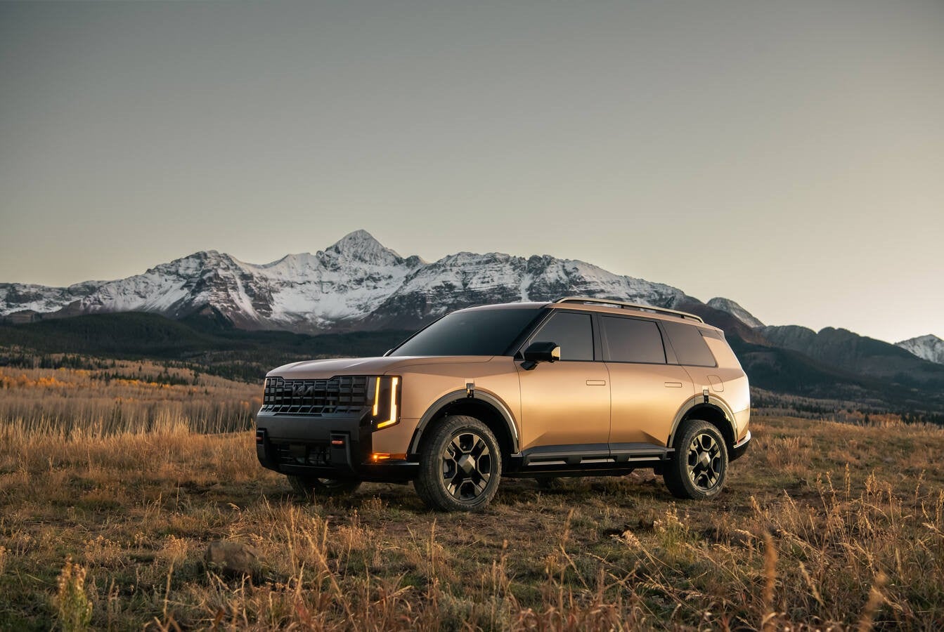 A brown American Made 2027 Kia Telluride in Phoenix is parked facing the viewer to the left on the grassy slope of a snow-covered mountain at dusk.