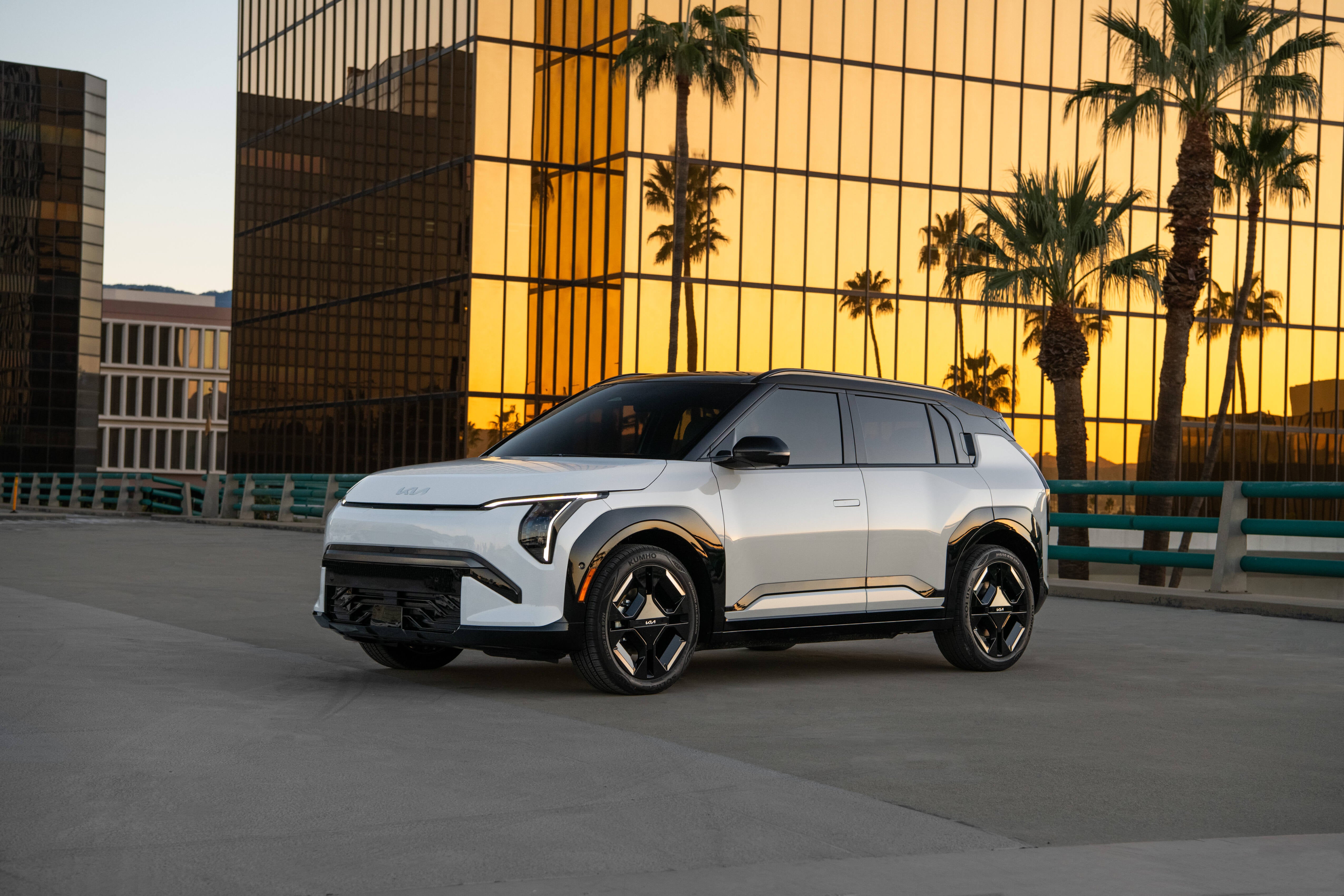 A white American Made 2027 Kia EV3 in Phoenix is parked facing the voewer to the left in an elevated city parking lot in front of palm trees and a golden glass building at sunset.
