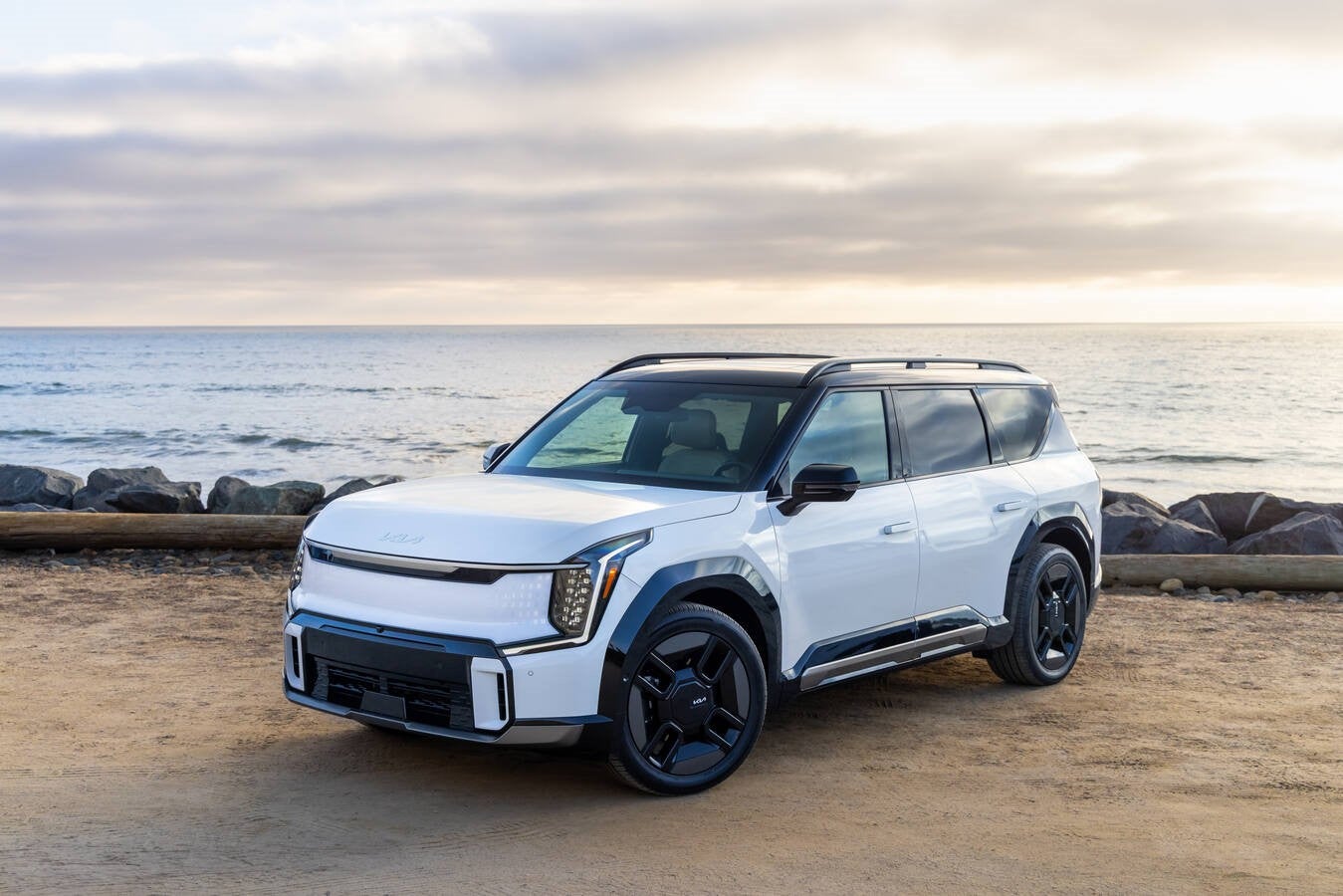 A white 2026 Kia EV9 in Peoria is parked facing the viewer to the left in a dirt parking lot in front of a large body of water.