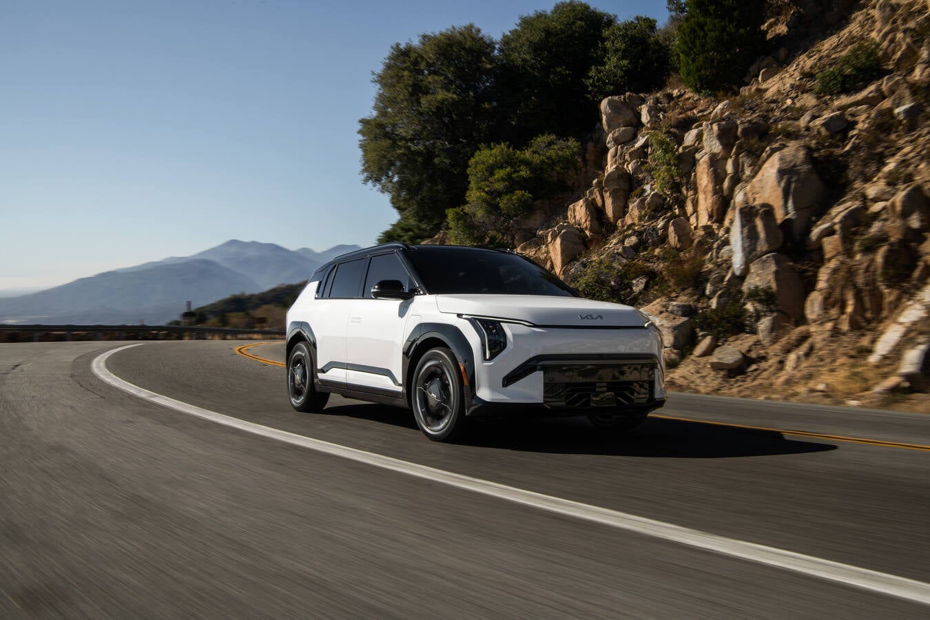 A white 2027 Kia EV3 in Peoria drives towards the viewer to the right on a curvy mountain road next to rocky cliffs.