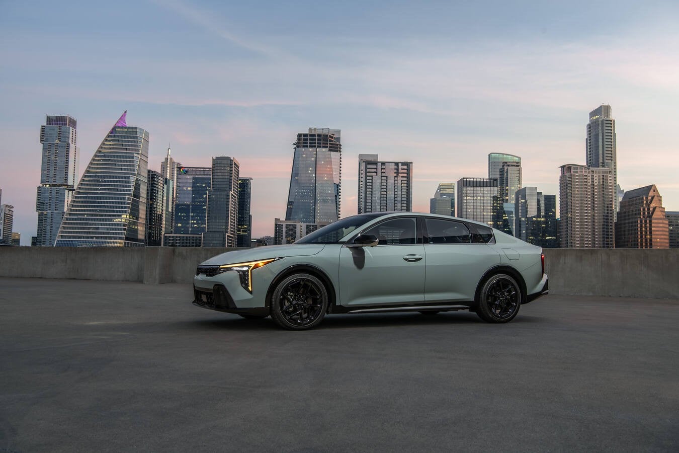 A light green 2026 Kia K4 in Phoenix is parked facing left on a concrete rooftop with a city skyline at dusk in the background.