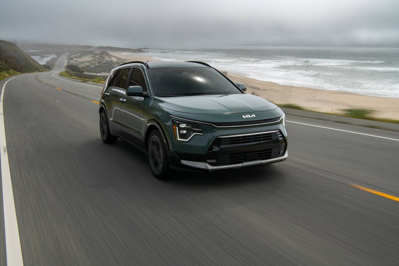 A dark green 2026 Kia Niro in Phoenix drives towards the viewer to the right up a coastal road with a beach in the background, seen from a high angle.