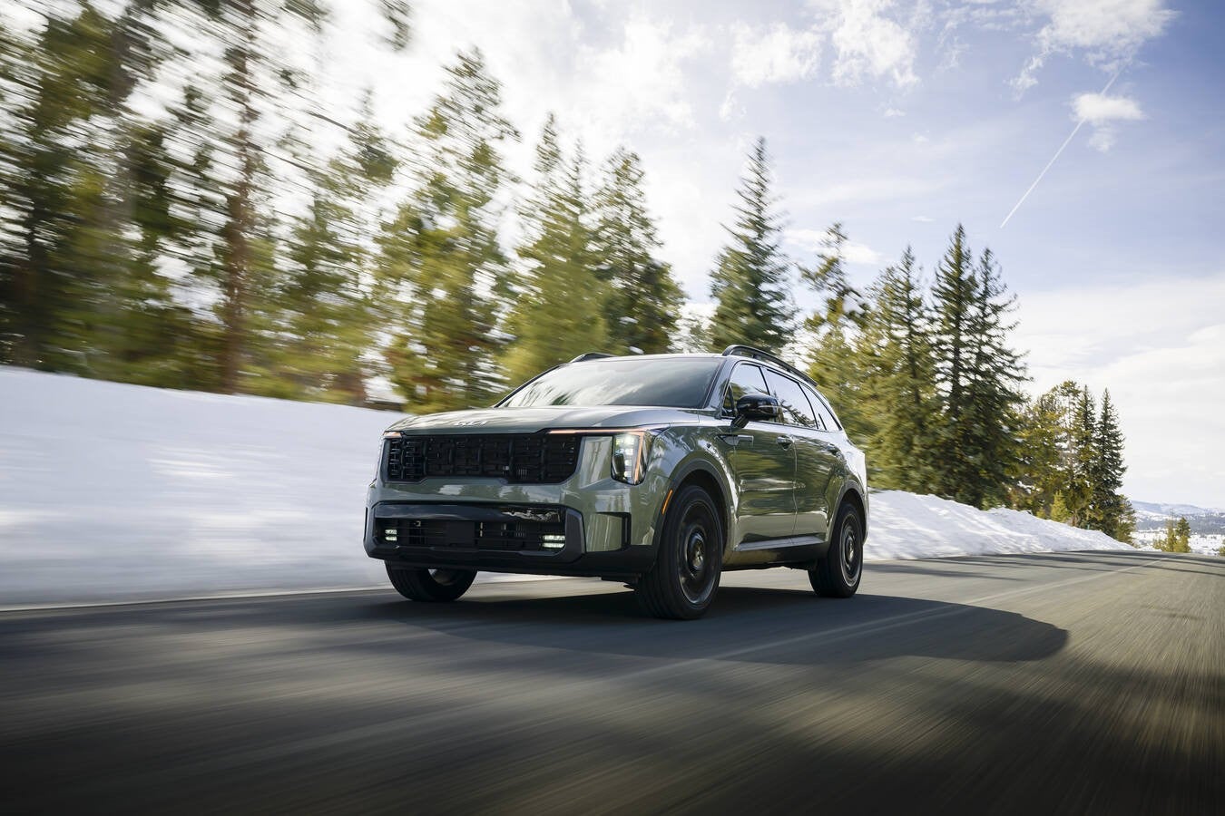 A green 2026 Kia Sorento in Peoria drives towards the viewer to the left on a two-lane road through a snowy landscape, seen from a low angle.