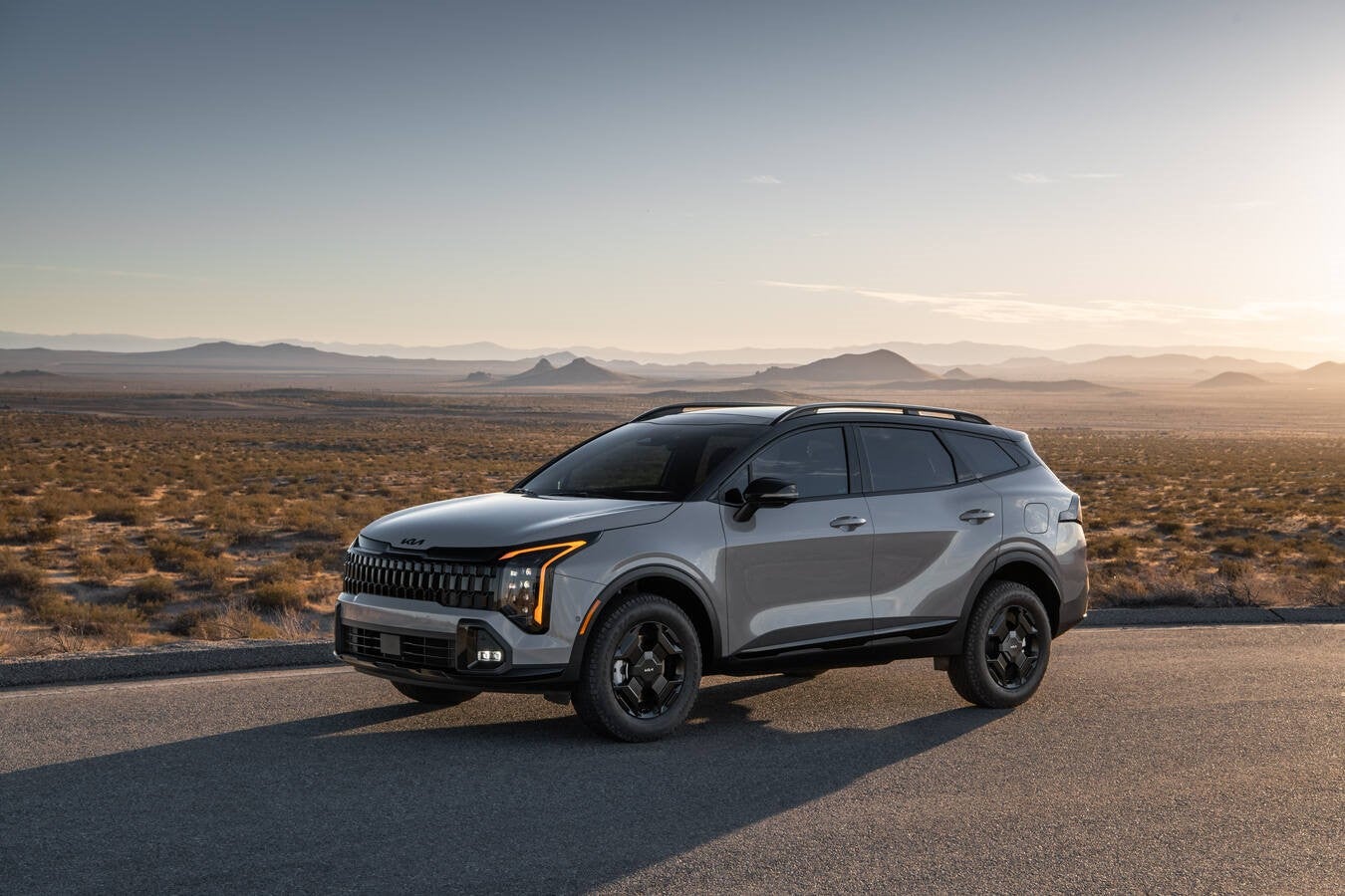 A grey 2026 Kia Sportage is parked facing the viewer to the left in front of a flat desert landscape with distant mountains in the background at sunset.