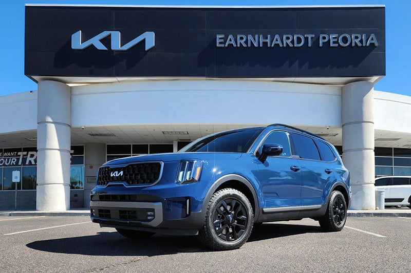 A blue Kia Telluride SUV is parked in front of the Earnhardt Peoria Kia dealership under a clear blue sky.