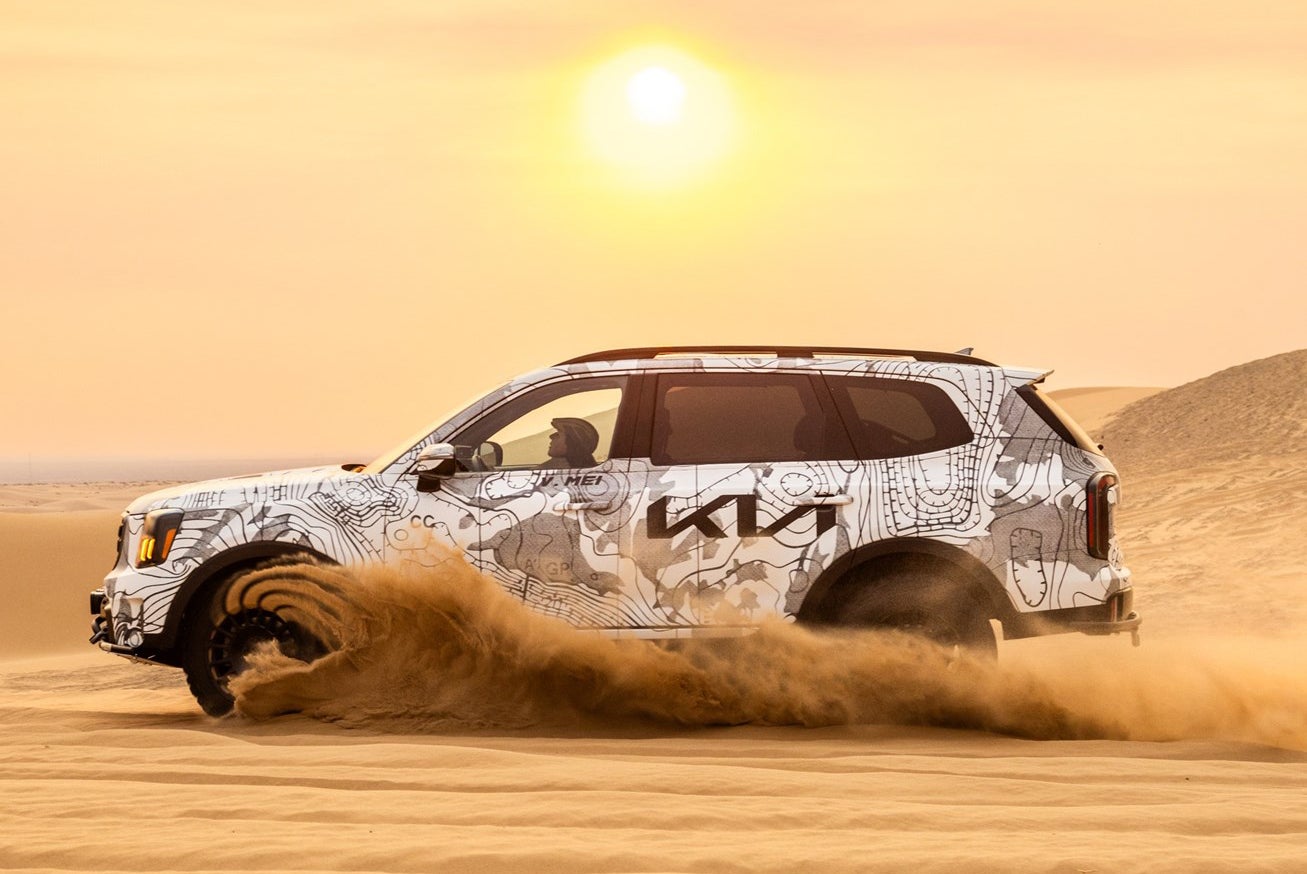Front view of a light gray Kia Telluride SUV driving on a coastal road with an ocean and hillside view.